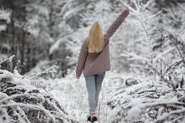 Милая модель девушка с длинными светлыми волосами в вязаном уютном кардигане ходит по зимнему заснеженному лесу — стоковое фото