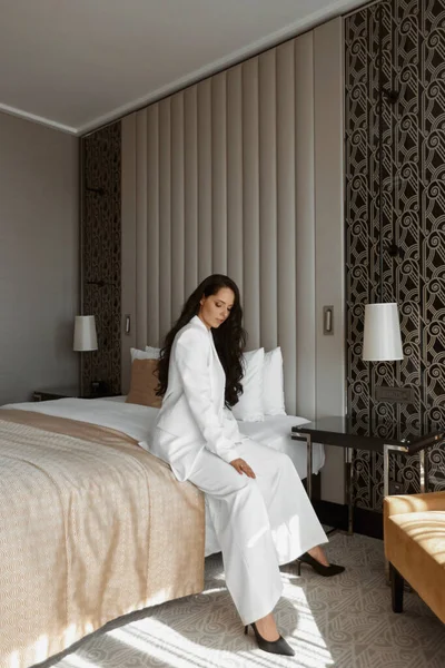 Young elegant lady in modish white suit and shoes sitting on the bed in interior — Stock Photo, Image