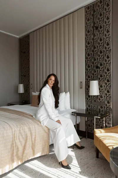 Pretty woman in a white trendy suit and high heels posing on the bed in modern interior — Stock Photo, Image