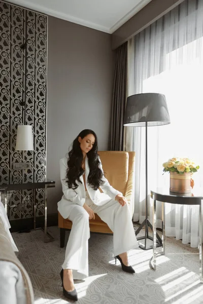 Vogue modeli beyaz şık takım elbise ve moda ayakkabılarla koltukta oturuyor.