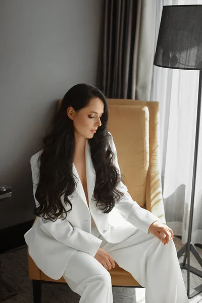 En fashionabel ung kvinna med perfekt långt svart hår och perfekt trendig makeup i en elegant vit kostym poserar i inredningen. Vacker brunett modell flicka i trendig poserar i en fåtölj — Stockfoto