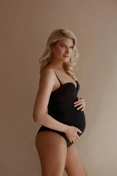 Sarışın, siyah elbiseli, bej arka planda poz veren çekici hamile kadın. — Stok fotoğraf