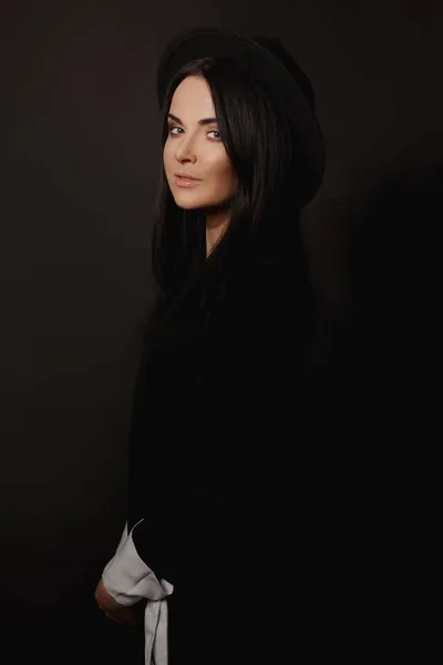 Adorale ung kvinna med trendig makeup bär svart trendig hatt och kort svart klänning poserar på den svarta bakgrunden — Stockfoto