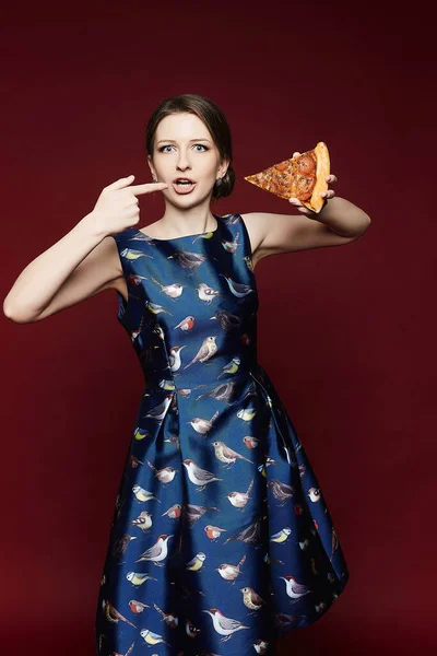Молодая модель женщина в модном синем платье держа кусок пиццы в руке и указывая на него, изолированные на красном фоне — стоковое фото