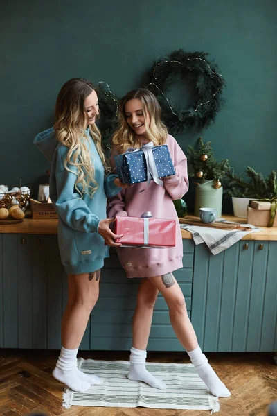 Duas belas mulheres namoradas em capuzes da moda dão umas às outras caixas de presente de Natal no interior decorado para feriados de Ano Novo. — Fotografia de Stock