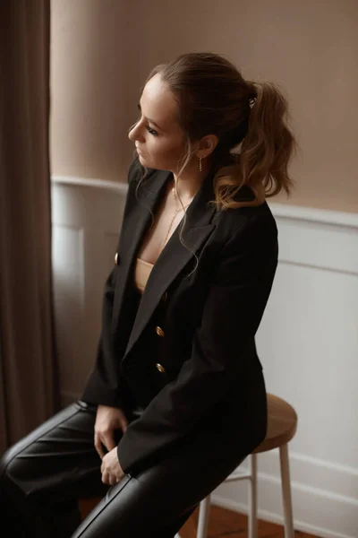 Moda makyajlı modaya uygun bir kadın modelin, şık bir spor ceket ve siyah pantolon giyen, oturma odasında poz veren bir portresi. — Stok fotoğraf