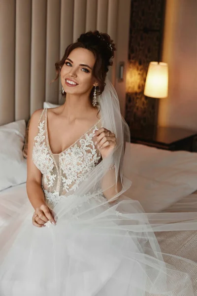 Adorabile giovane sposa con acconciatura elegante indossa abito alla moda e orecchini seduti sul letto — Foto Stock