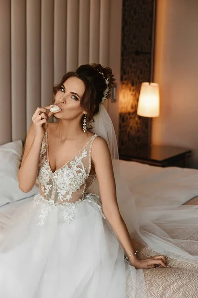 Splendida giovane sposa in un abito da sposa alla moda godendo gustoso dessert mentre seduto sul letto — Foto Stock