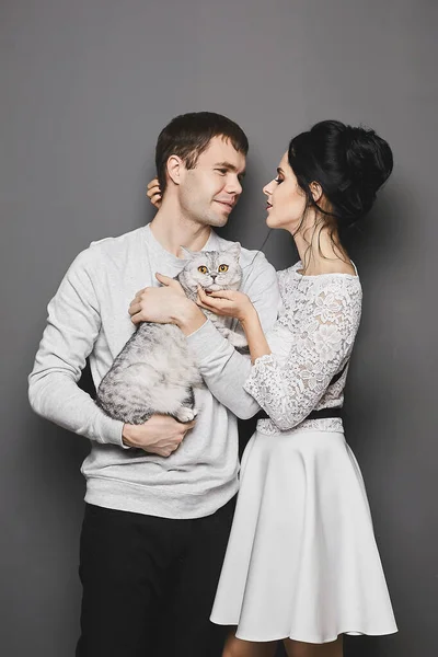 Sweet couple of lovers, a young gorgeous woman in dress and handsome guy posing with their cute cat on the grey background — 图库照片