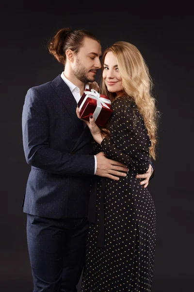 A handsome guy in a stylish suit giving a red gift box to his charming girlfriend wearing a trendy dress. Valentines day concept — Fotografia de Stock
