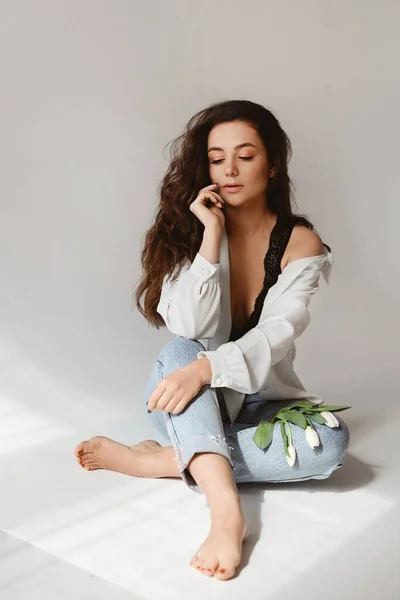 Beautifiul young woman with nude makeup wearing jeans and unbuttoned blouse posing with a sprig of flowers on the floor — 图库照片
