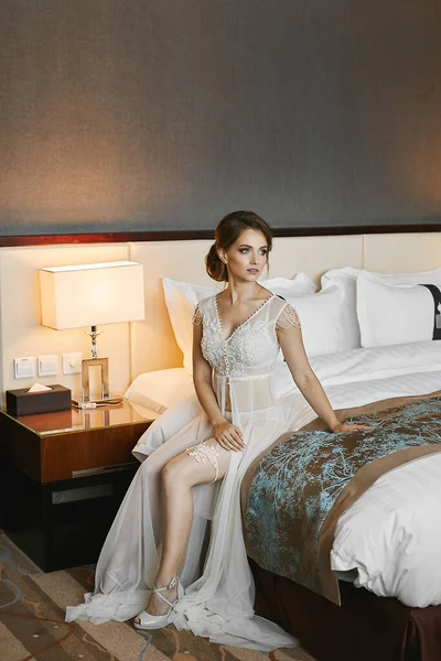 Joven novia seductora con maquillaje perfecto y peinado en vestido de tocador transparente blanco de tul y encaje sentado en una cama — Foto de Stock