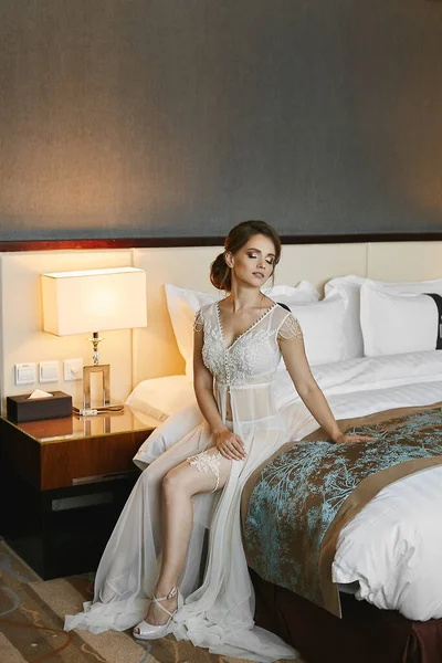 Encantadora modelo mujer con maquillaje perfecto y peinado modesto en vestido de boudoir transparente blanco de tul y encaje sentado en una cama — Foto de Stock