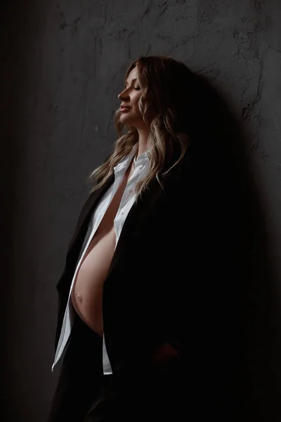 Een prachtige zwangere, jonge blonde vrouw met een ongeknoopt wit shirt en blazer blijft met gesloten ogen naast de zwarte muur. Naakte buik. Donkere achtergrond. Lifestyle, zwangerschap, gezondheid — Stockfoto