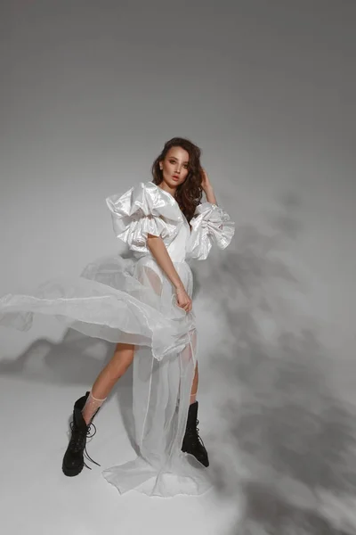 Hög mode skott av en ung modell kvinna i en trendig fladdrande vit klänning och modisk svart läder stövlar på den grå bakgrunden. Modetrender. Ung brud i trendig brudklänning — Stockfoto