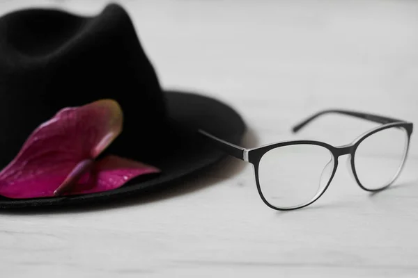 Schwarzer schicker Hut mit pinkfarbener Blume liegt neben modischen Brillen auf einem weißen Holztisch Stockbild