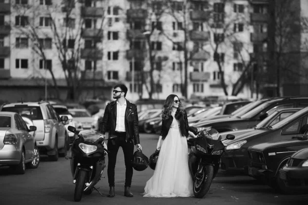 Stylish bride and bearded groom stand beside sports motorcycles on the urban street. A beautiful couple of newlyweds posing with their motorbikes — Stock Photo, Image