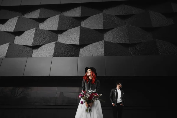 Noivo elegante em jaqueta de couro e óculos de sol e noiva de cabelos vermelhos na moda em um vestido branco, chapéu preto e jaqueta de couro, posando juntos no fundo urbano preto. Conceito de um dia de casamento. — Fotografia de Stock