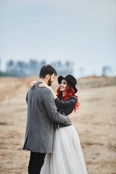 Una coppia di sposi alla moda in posa all'aperto. Elegante uomo barbuto e la sua ragazza dai capelli rossi in un abito da sposa e giacca di pelle — Foto Stock