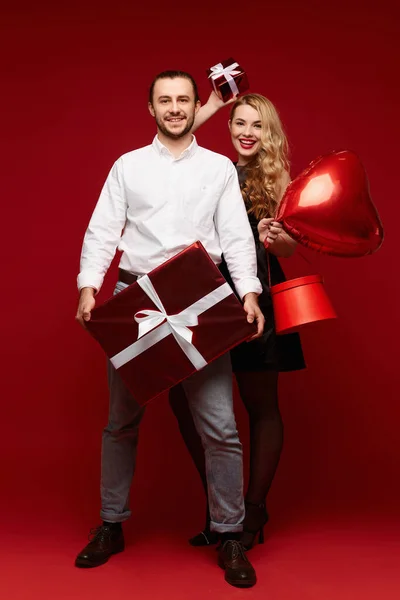 Um lindo casal heterossexual posando com caixas de presente e um balão vermelho na forma de um coração, uma mulher loira bonita e um jovem bonito posando no fundo vermelho Fotografias De Stock Royalty-Free