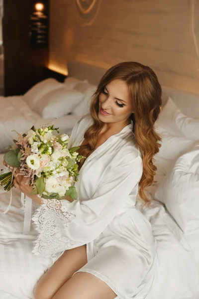 Glückliche junge Frau im Morgenmantel genießt mit einem Brautstrauß auf dem Bett lizenzfreie Stockbilder