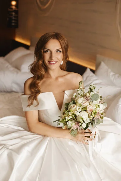 Glückliche junge Braut im modischen Hochzeitskleid sitzt mit einem stilvollen Strauß auf dem Bett Stockfoto
