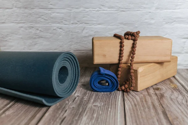 Closeup of two wooden yoga blocks, belt, rudraksha rosary and blue yoga mat on the wooden floor.