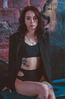 Dövmeli bir kız ceketli ve iç çamaşırlı bir şifoniyerin üzerinde oturuyor.