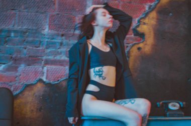 Dövmeli bir kız ceketli ve iç çamaşırlı bir şifoniyerin üzerinde oturuyor.