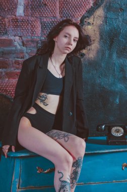 Dövmeli bir kız ceketli ve iç çamaşırlı bir şifoniyerin üzerinde oturuyor.