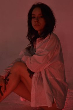 Beyaz tişörtlü erotik kız pembe neonlu yerde oturuyor.