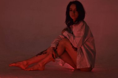 Beyaz tişörtlü erotik kız pembe neonlu yerde oturuyor.