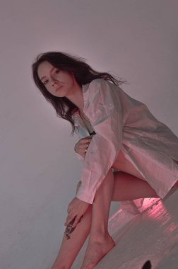 Beyaz tişörtlü erotik kız pembe neonlu yerde oturuyor.