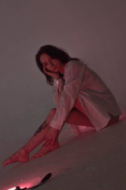 Beyaz tişörtlü erotik kız pembe neonlu yerde oturuyor.