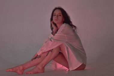 Beyaz tişörtlü erotik kız pembe neonlu yerde oturuyor.