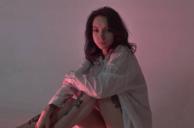 Dövmeli bir kız ceketli ve iç çamaşırlı bir şifoniyerin üzerinde oturuyor.