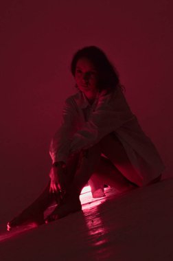 Beyaz tişörtlü erotik kız pembe neonlu yerde oturuyor.