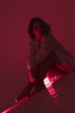 Beyaz tişörtlü erotik kız pembe neonlu yerde oturuyor.