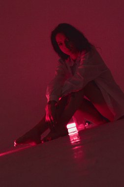 Beyaz tişörtlü erotik kız pembe neonlu yerde oturuyor.