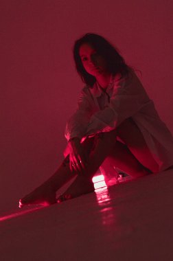 Beyaz tişörtlü erotik kız pembe neonlu yerde oturuyor.