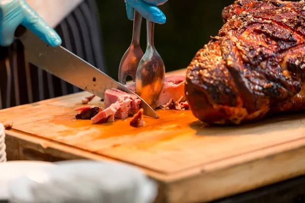 Grill restaurant kitchen. Chef in Blue cooking gloves using knife to cut smoked pork. Roast pork neck. Roasted shoulder of pork on a cutting board.