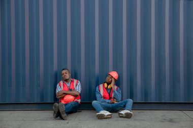 Amerikalı Afrikalı teknisyenler kargo konteynırının yanında oturuyorlar ve yorgun, uykulu ya da işini kaybetmiş görünüyorlar. Fabrika işçisi kargo konteynırının yanında oturur ve yorgun görünür ve işini kaybeder..