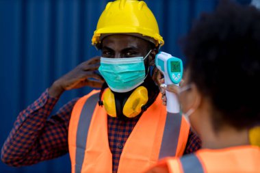 Temperature screening and medical check at Container cargo site. Worker in face mask on work site in covid-19 outbreak. Foreman man  with thermometer at site. Social distancing. Coronavirus prevention