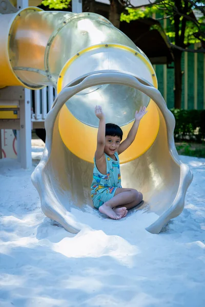 Boy playing on children\'s slides. Child Playing slider at outdoor summer playground. Child playing slider on sand ground. Kid sitting on slider of kid play ground in public park.