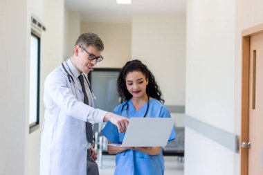 Üniformalı klinisyenler ayakta dikilip bilgisayar monitörlerine bakıyor, tıbbi belgeleri tartışıyor ve danışmanlık yapıyorlar. Doktor ve hemşire tıp bürosunda dizüstü bilgisayarla çalışıyorlar. Dizüstü bilgisayarla profesyonel tıbbi işler.