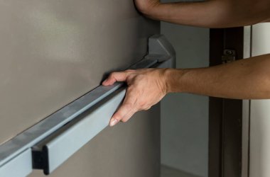 Hand is Opening the emergency fire exit door. Emergency fire exit door. Closed up latch and rusty door handle of emergency exit. Push bar and rail for panic exit. Open one way door.