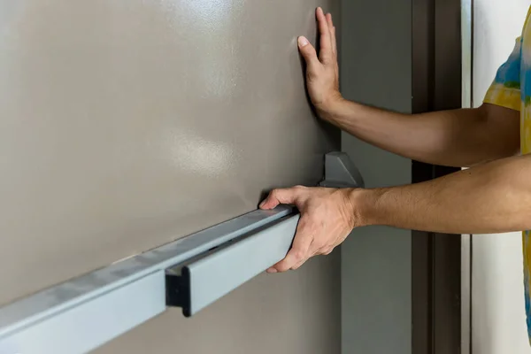 Hand is Opening the emergency fire exit door. Emergency fire exit door. Closed up latch and rusty door handle of emergency exit. Push bar and rail for panic exit. Open one way door.