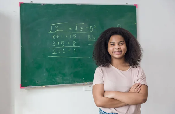 Girl is standing in a classroom with a green chalkboard covered with formulas and figures. Kid write on chalk board.