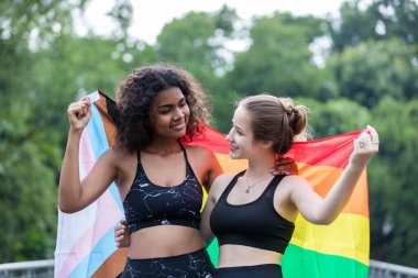 Two  young women standing with a pride rainbow flag. couple hugging at outdoor. LGBT rainbow flag.