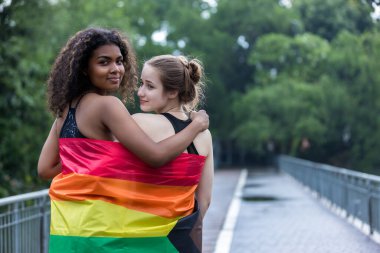 Two  young women standing with a pride rainbow flag. couple hugging at outdoor. LGBT rainbow flag.